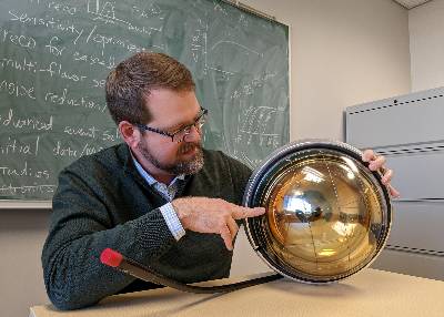 MSU Professor Tyce DeYoung sits in front of a green chalkboard covered with notes, graphs and equations. He’s holding a metal and glass gold orb, which is a digital optical module that IceCube uses to detect neutrinos.