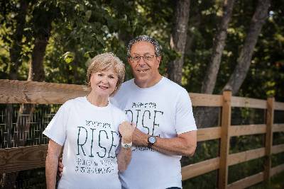 A photo of Sue and Gary Farha standing outside with a fence and wooded area in the background. They are both wearing white RISE t-shirts.