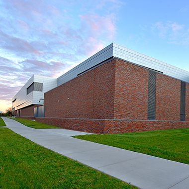 The metal-capped brick exterior of the FRIB building sits behind a bright green lawn and under a blue sky, dusted with white clouds starting to take on orange and purple hues of dusk.
