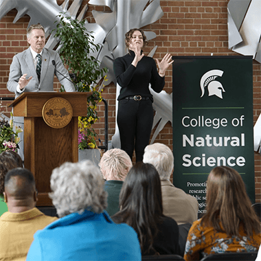 MSU President Kevin M. Guskiewicz stands in front of a lectern on stage with an ASL interpreter and a sign that says “College of Natural Science.”